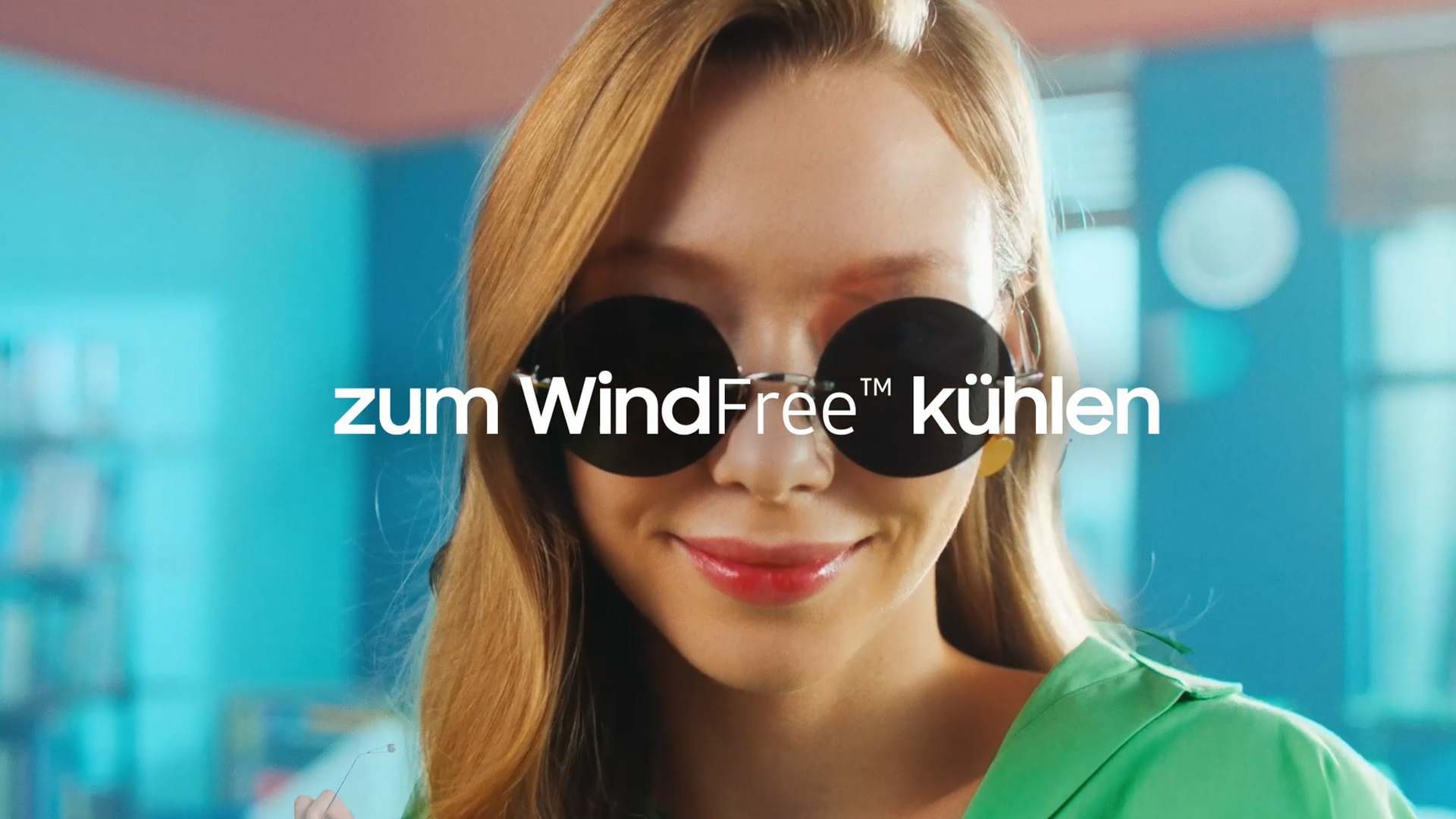 MTF Shop Samsung Klimaanlage Wärmepumpe Österreich | Eine lächelnde Frau mit Sonnenbrille vor einem türkisfarbenen Hintergrund. Der Text im Bild „zum WindFree kühlen“ weist auf ein Video hin, das die Vorteile der zugfreien Samsung WindFree Kühltechnologie für maximalen Komfort erklärt.